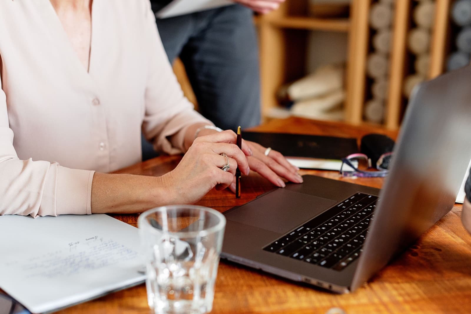 Close-up of a woman's hands holding a pen at a laptop beside handwritten notes and a glass of water