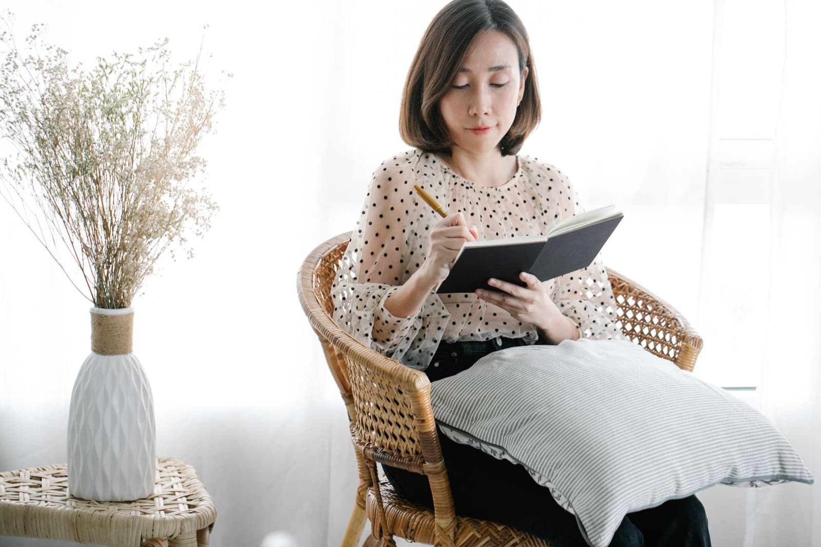 Woman writing in a journal while sitting in a wicker chair by a sunlit window, with a vase of dried flowers nearby