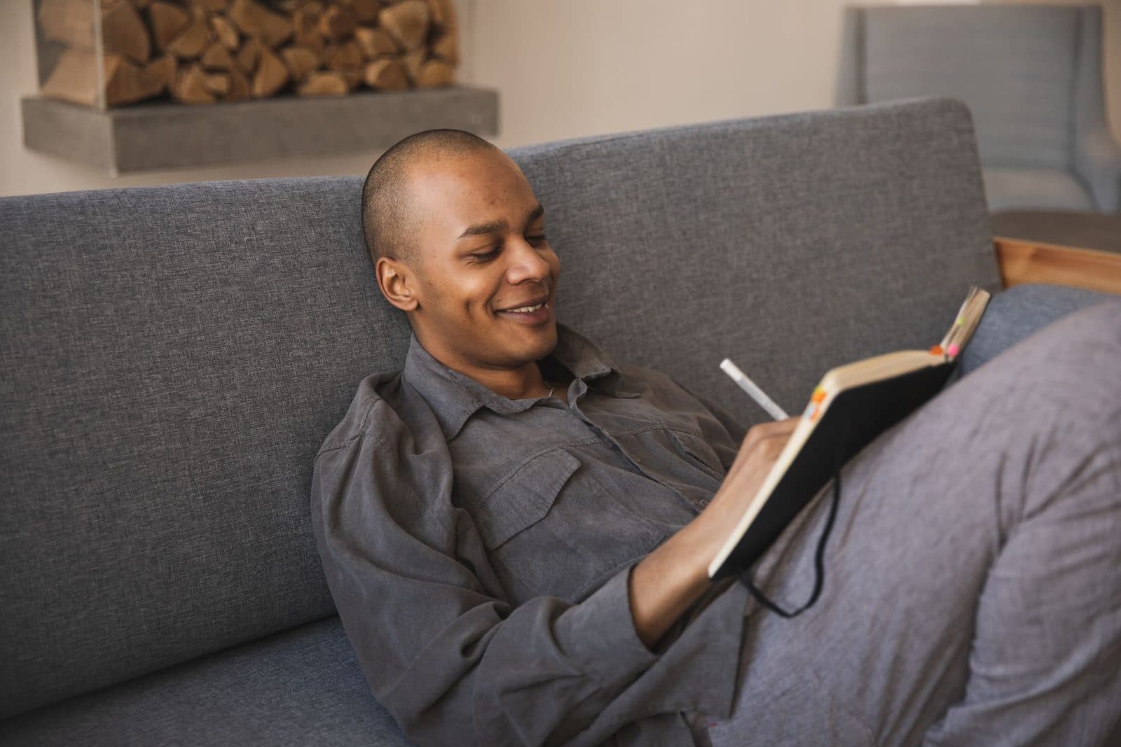 Smiling man in a gray shirt writing in a notebook while relaxing on a gray sofa at home