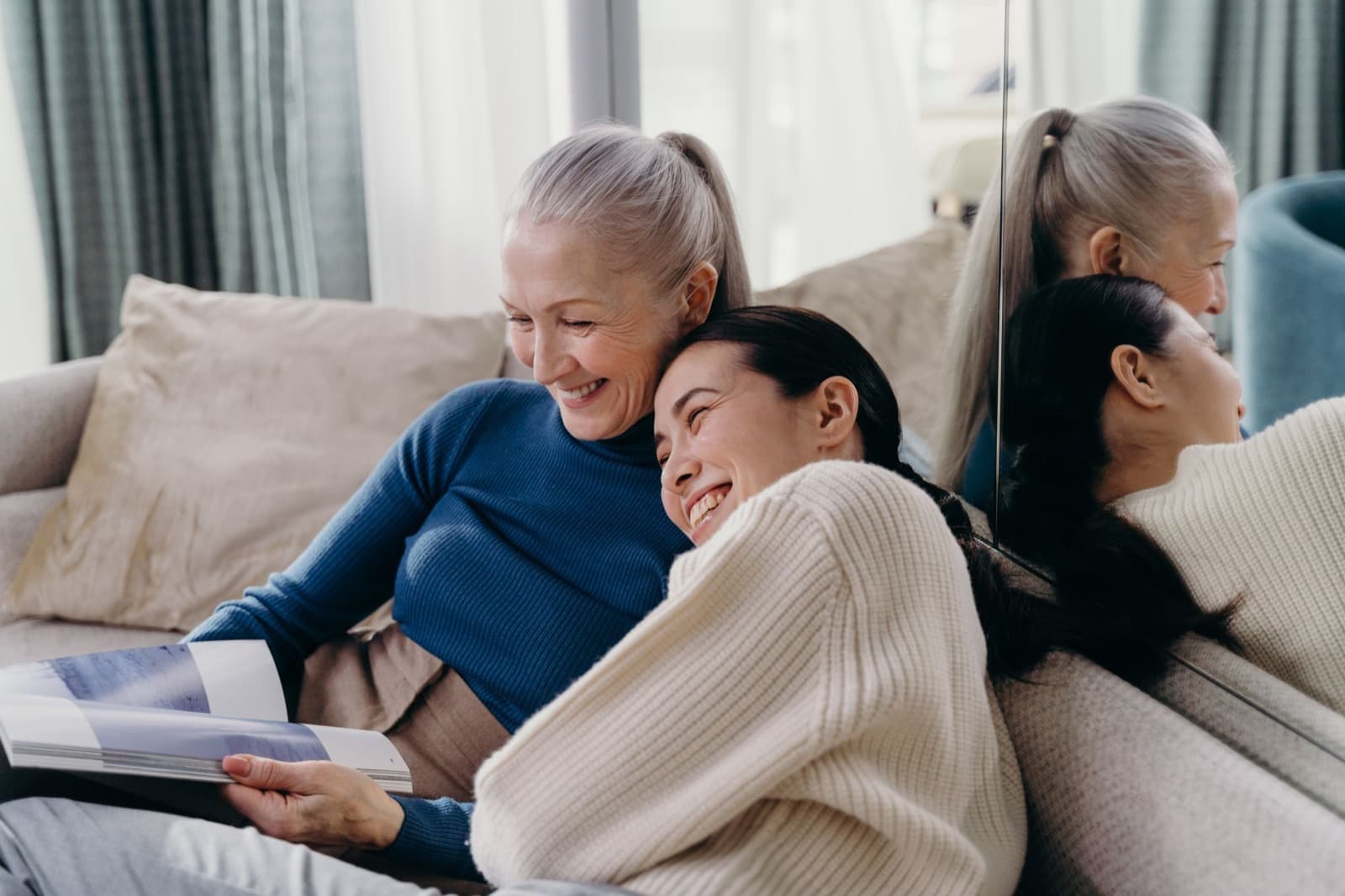 Older woman with a gray ponytail and a younger woman laughing together on a sofa while looking through a magazine