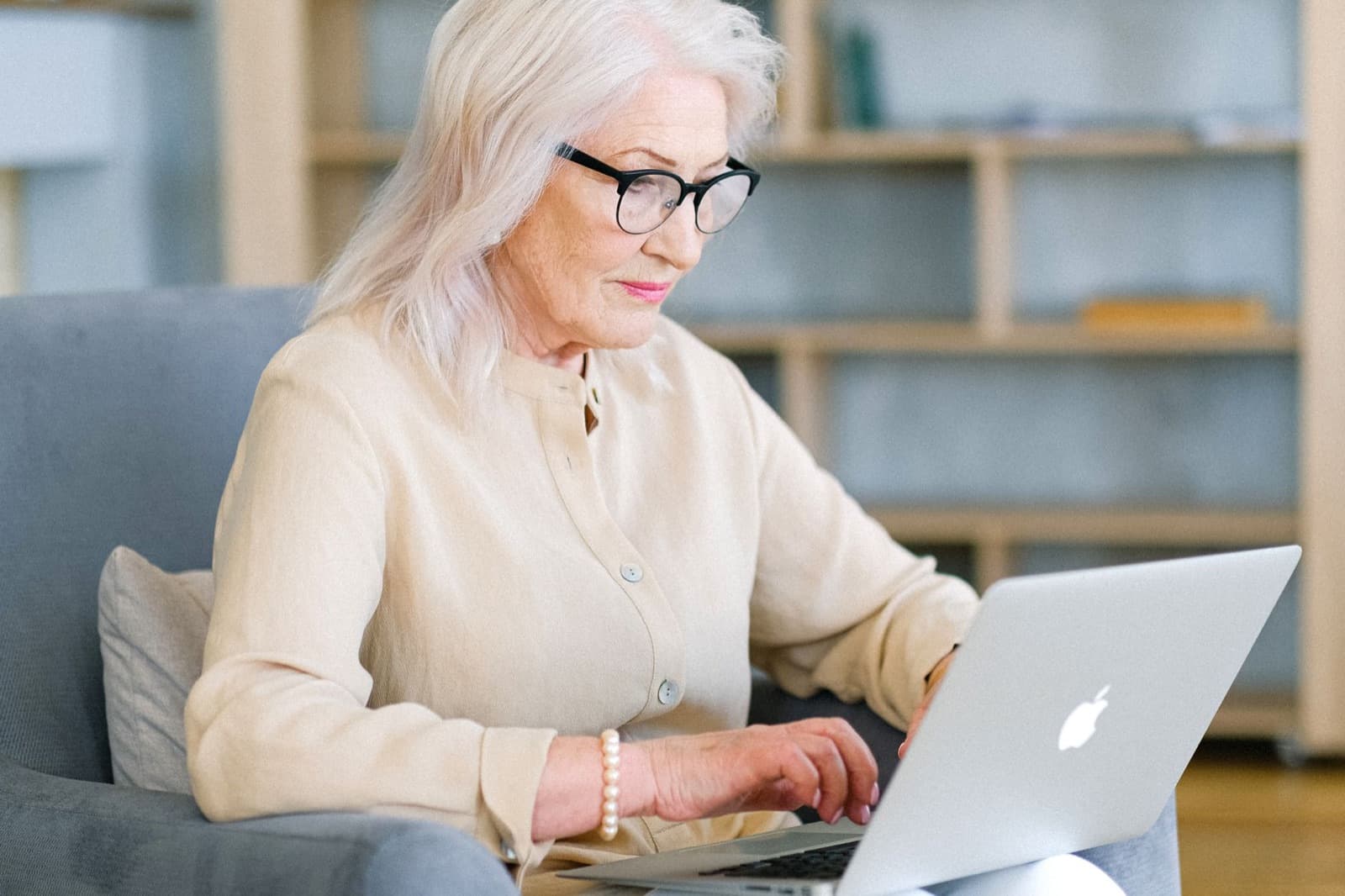 Older woman with silver hair and glasses using a laptop while seated in a gray armchair at home