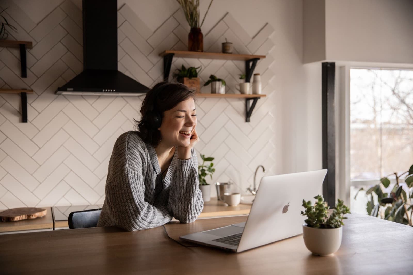Smiling woman wearing headphones talking to someone on a laptop at a wooden kitchen table